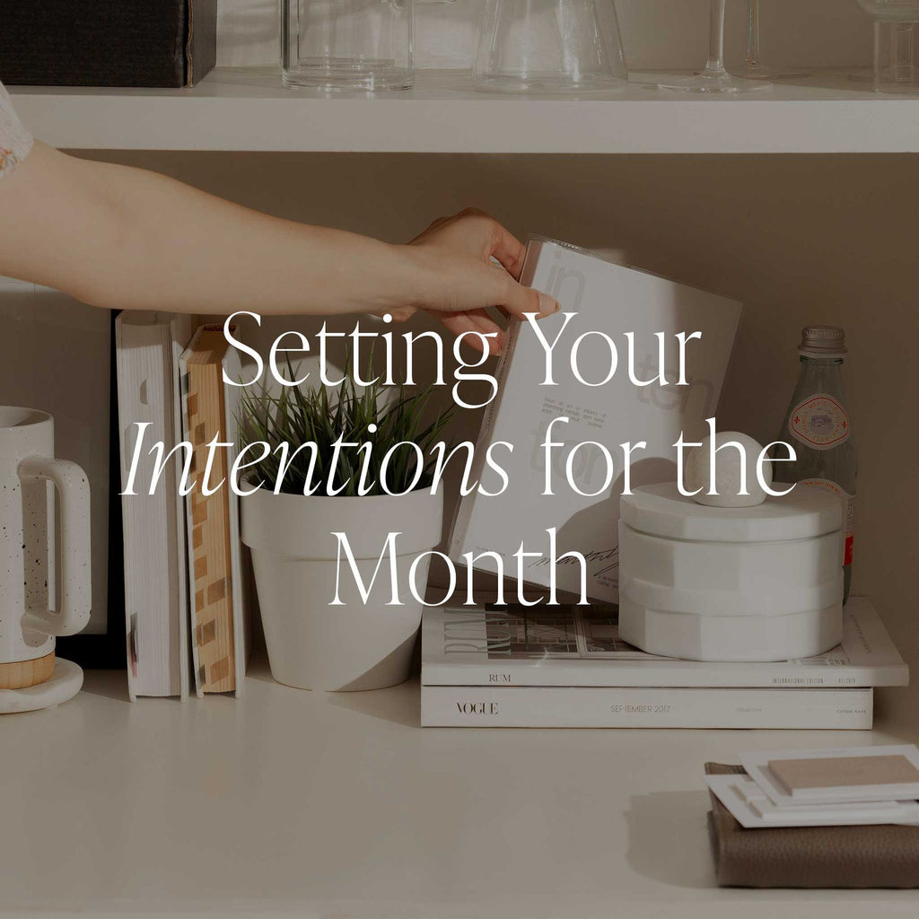 An image of a hand reaching for a notebook on a white shelf filled with books, indoor cactus, and a white decorative ceramic jar. It has a text overlay that reads: "Setting Your Intentions for the Month." 