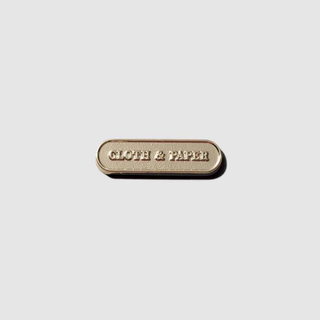 Gold pin displayed on a neutral background. Pin is pill shaped and has the Cloth and Paper logo inside of it.