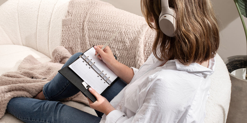 A person is sitting on a sofa, working in their personal planner with with 2024 planner inserts.  There is a pen in their right hand and they are listening to music on their headphones.