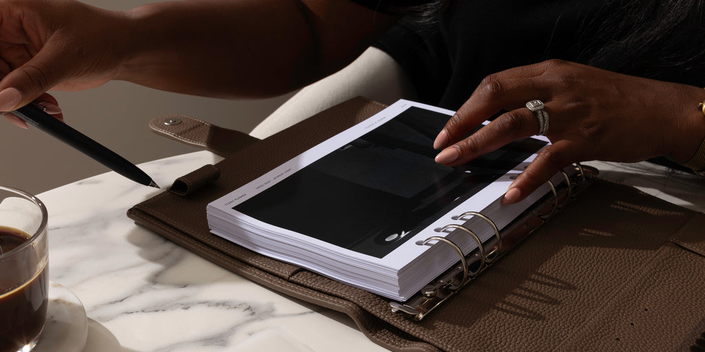 A left hand is removing a pen from a leather pen loop while a right hand is resting on a cover page from a set of 2024 inserts that are inside the leather planner.