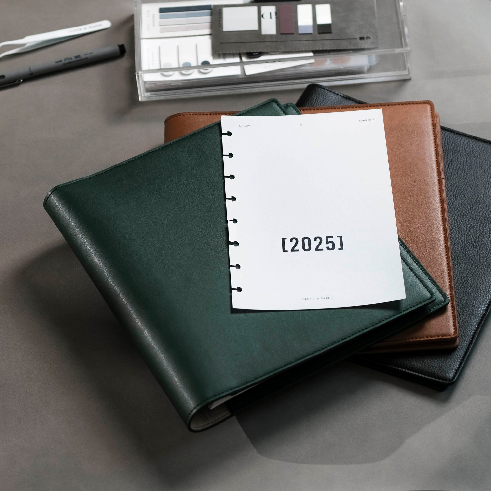 A few Cactus Vegan Leather Folios are sitting on a dark gray desk with other surrounding planner accessories.