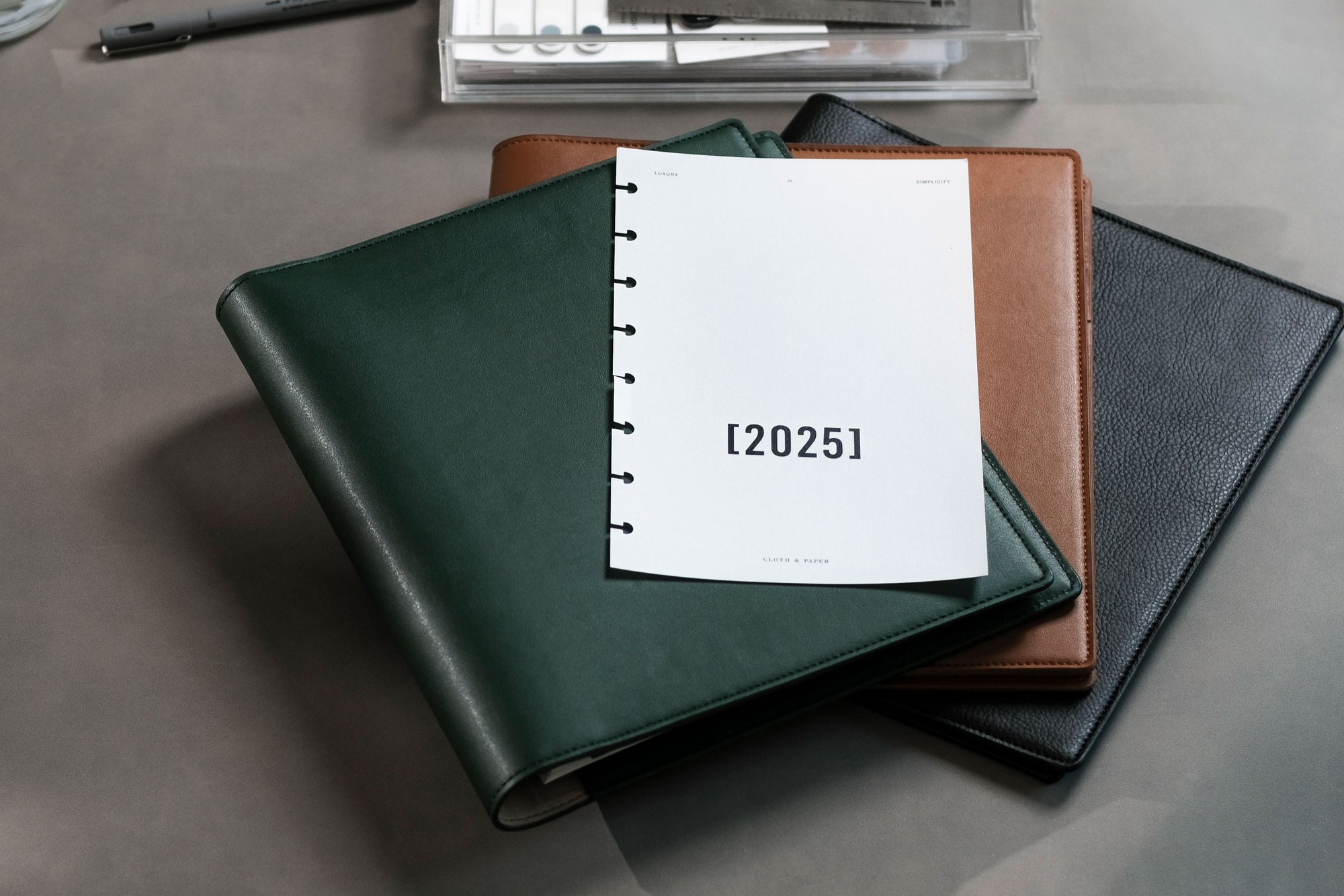 A few Cactus Vegan Leather Folios are sitting on a dark gray desk with other surrounding planner accessories.