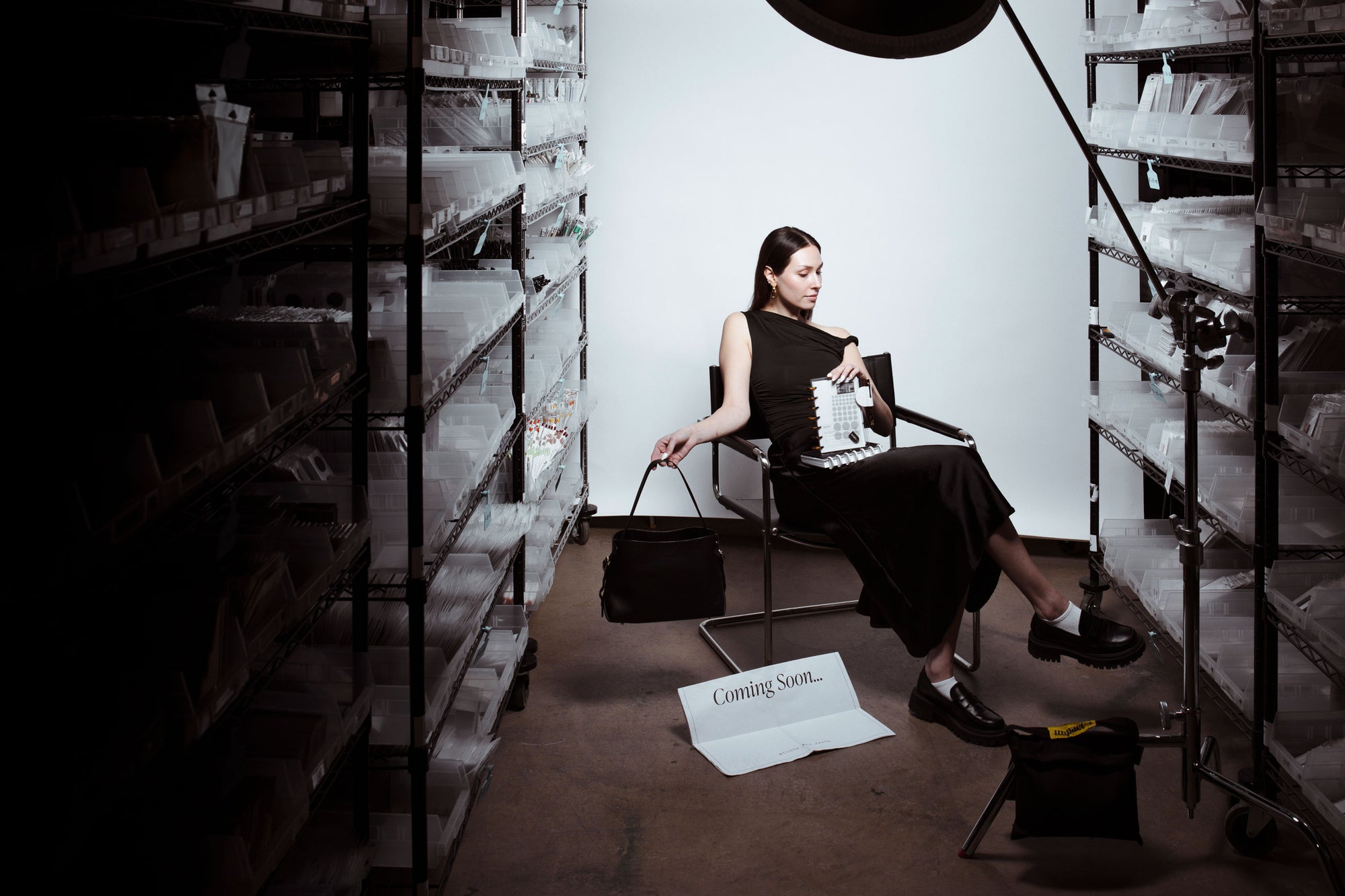 A model is sitting in a chair beneath a spot light holding a couple assembled discbound planners in her lap and a leather Valencia Bag in her other hand. There are shelves on the left and right of her of planner essentials.