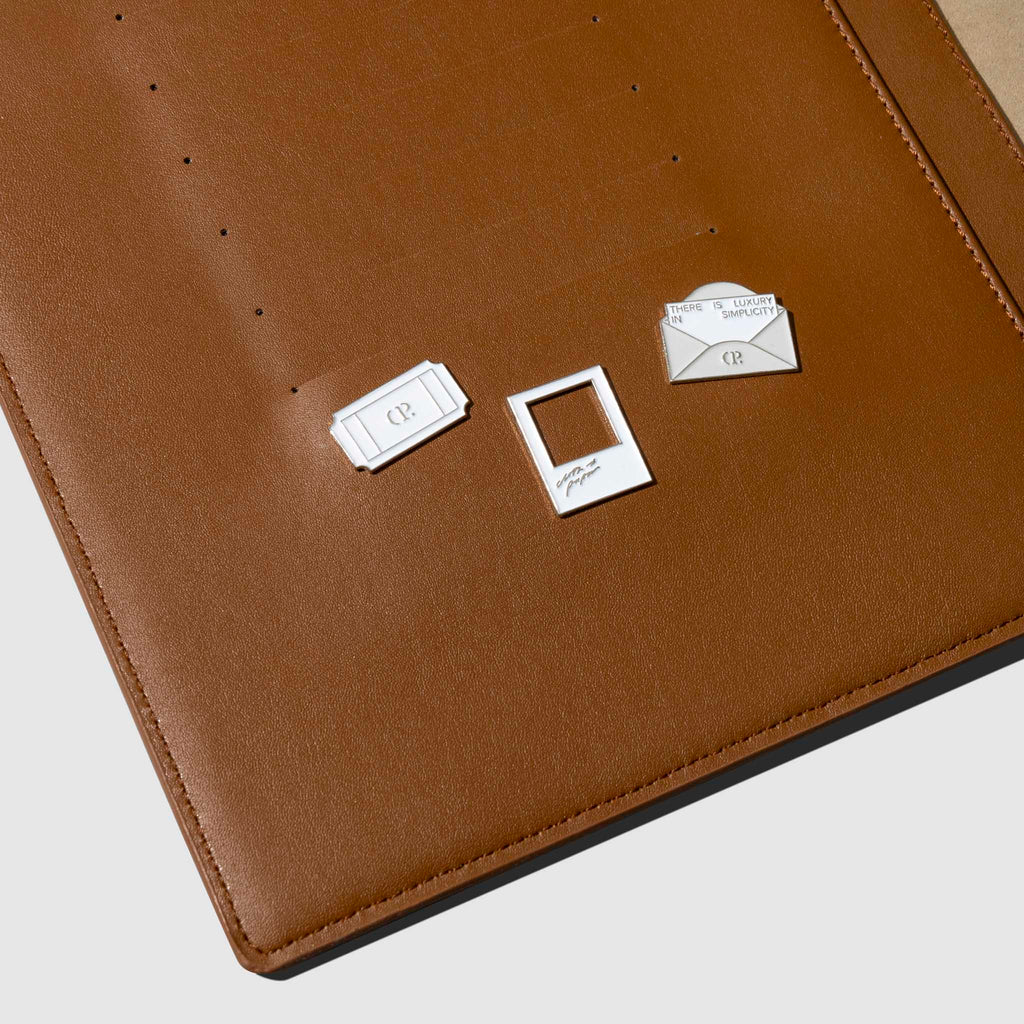 Set of 3 enamel pins displayed in use inside a brown leather planner.