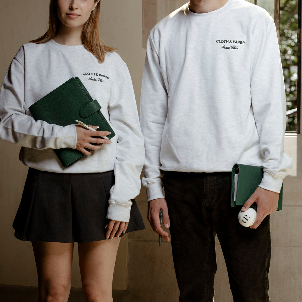 Two models, male and female, pictured from the chin down wearing Social Club sweatshirts. Both models are holding green leather agendas.