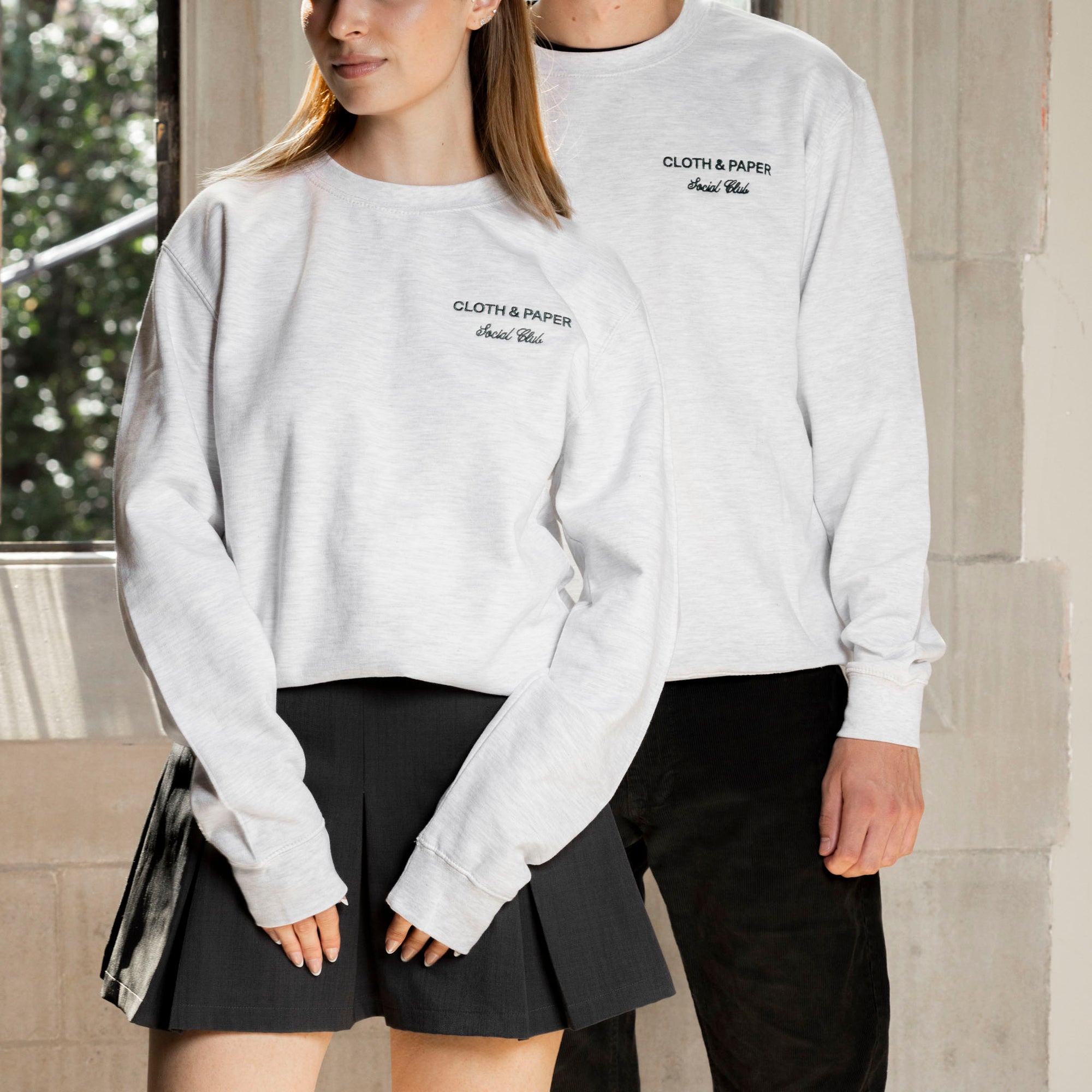 A male and female model are standing in a brightly lit area, each wearing a sweatshirt with black bottoms. 