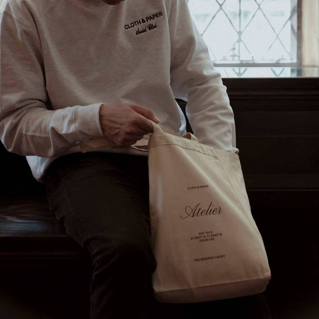 A male model, shown seated, reaches into a Cloth and Paper tote bag. He is wearing a social club crewneck.