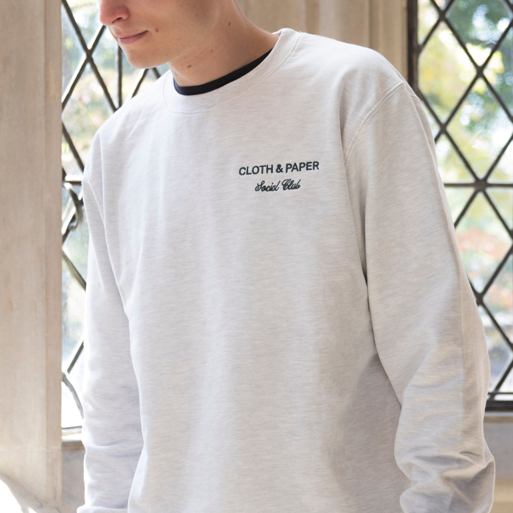 A male model is shown, from the chin down, wearing a Social Club crewneck. A brightly lit window is behind him.