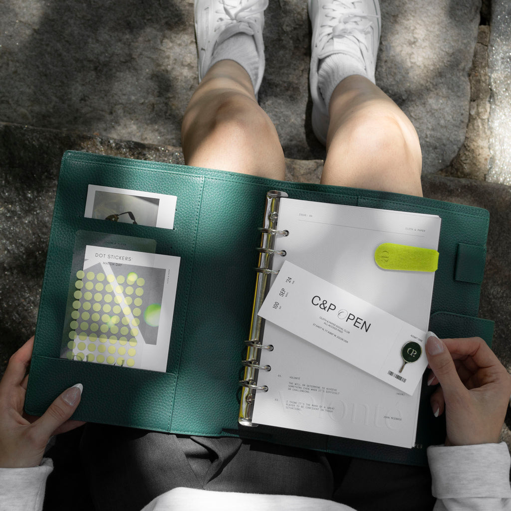 A female model, pictured from the top down, shown holding a green leather agenda in her lap. The agenda has a dashboard, clip, enamel pin, and sticker set inside of it.