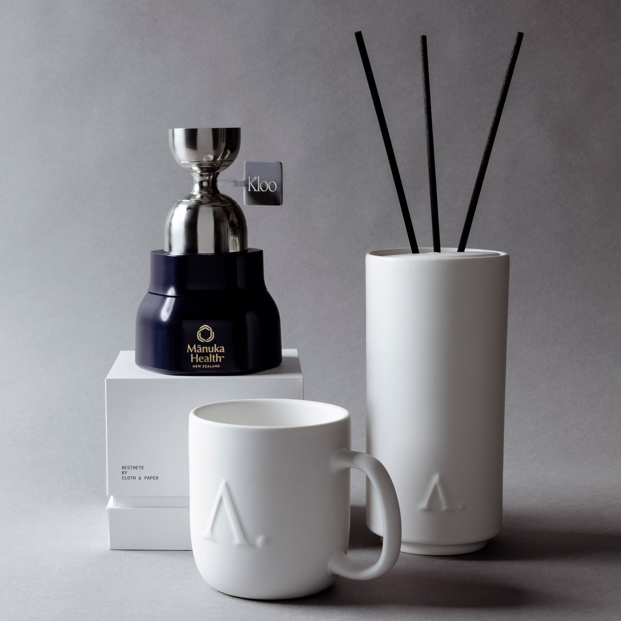 Aesthete Luxury Lifestyle Subscription Box against a gray background.  Mānuka Health Honey, Aesthete Ceramic Coffee Mug, and Basilica Diffuser.