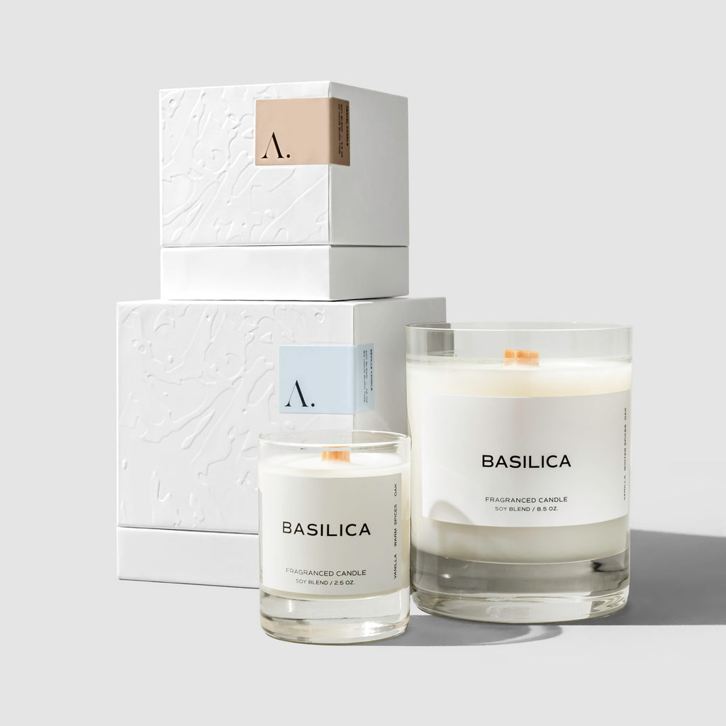 Full sized Basilica candle shown next to a travel size Basilica candle. Both of their boxes are shown in the background, stacked on top of each other.