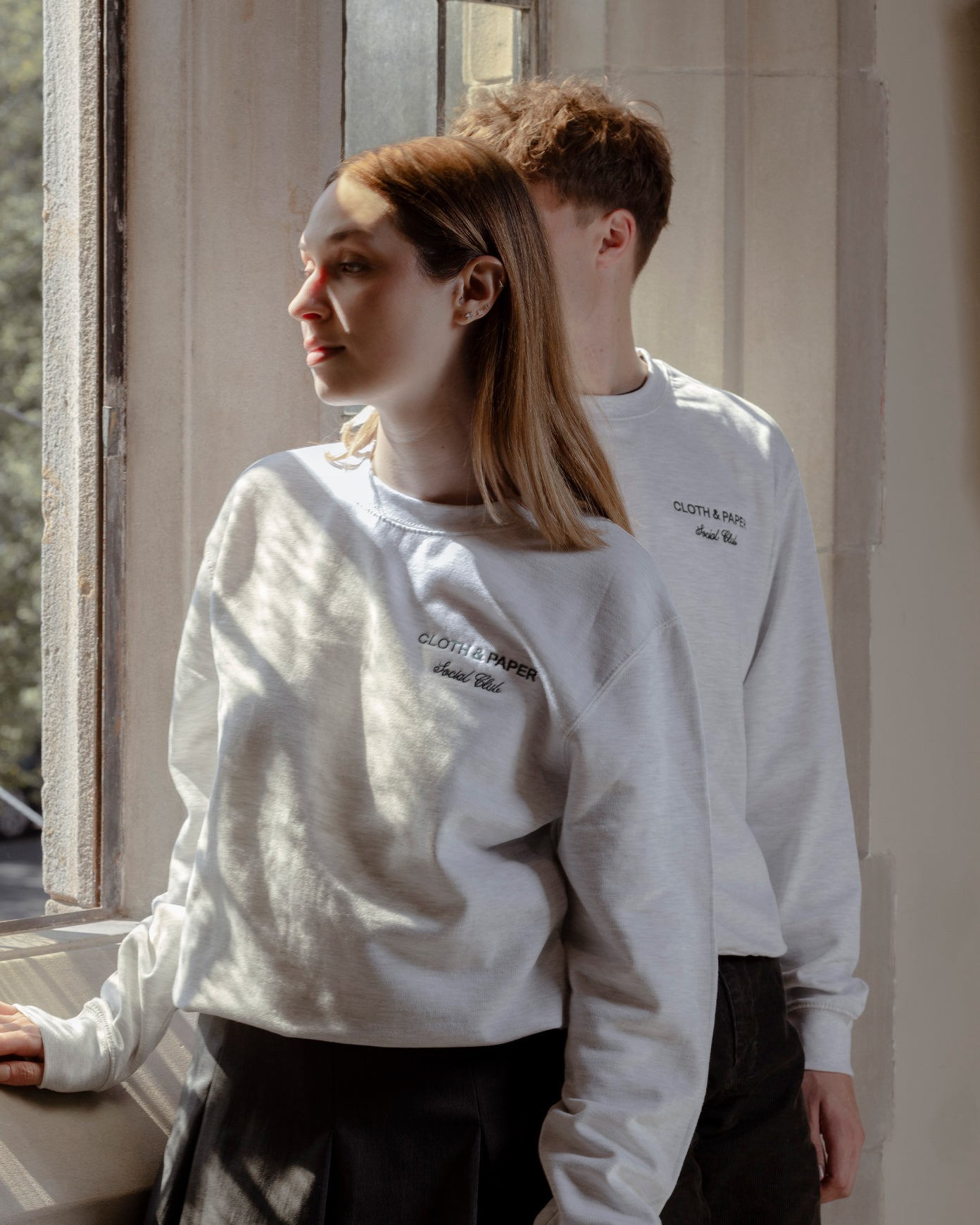 Two models are wearing Social Club Crewnecks.  They are standing next to an open window with sunshine coming through.  The green embroidery reads Cloth & Paper Social Club.