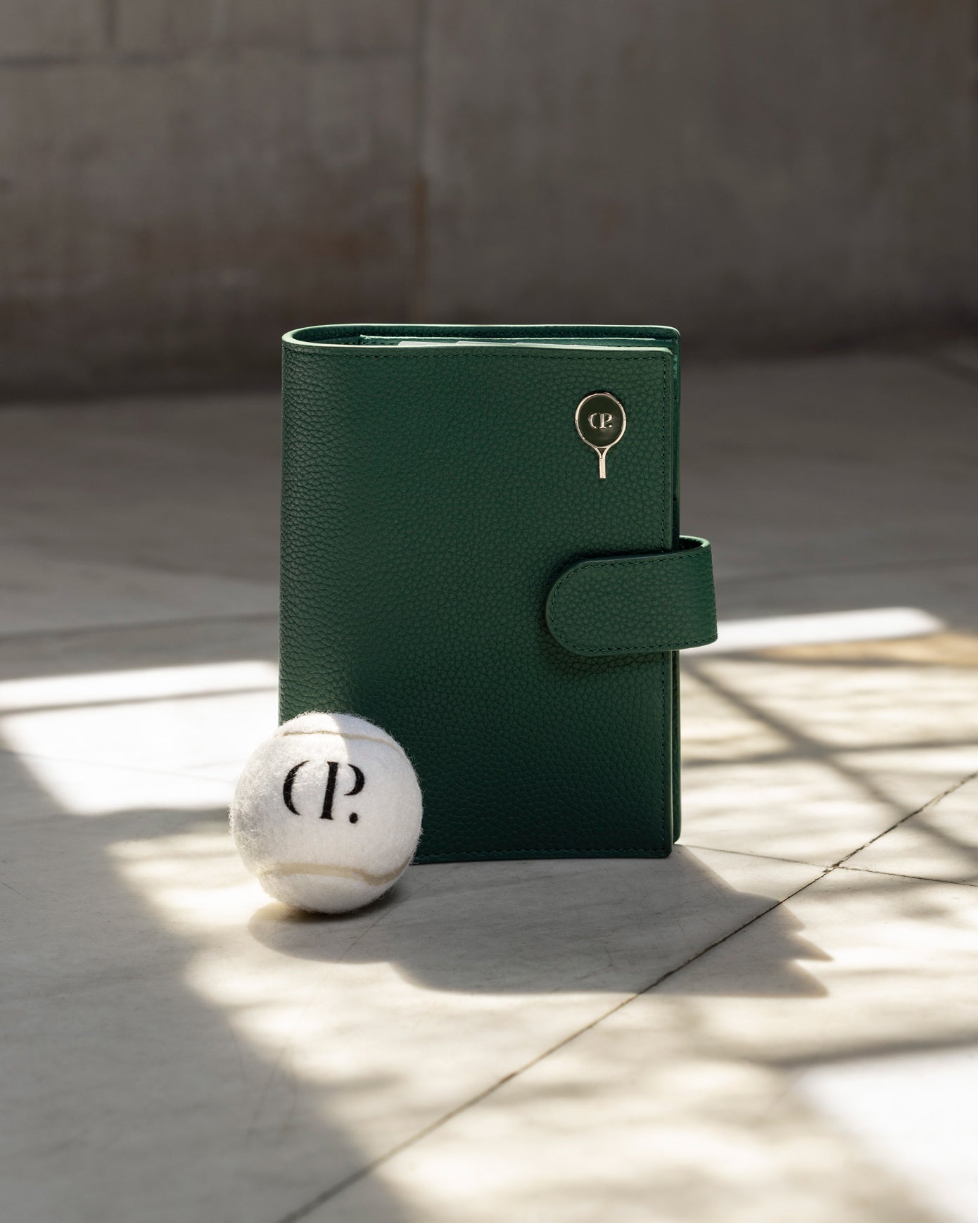 A 6-Ring Leather Agenda in the share of green is standing up in the sunlight.  There is a tennis ball with the CP logo and an Enamel Pin Racket present as well.