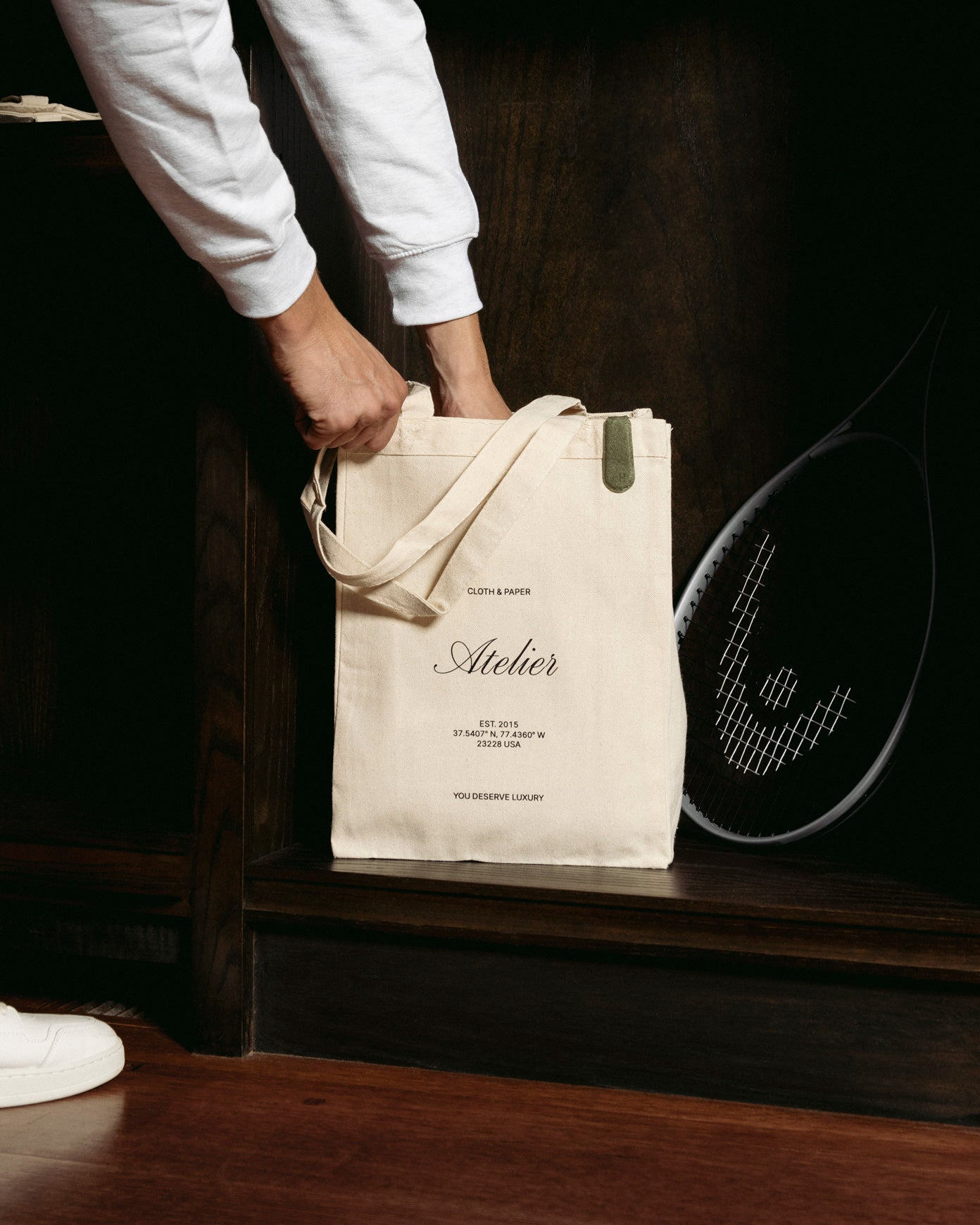 A model is reaching into the Atelier Tote Bag.  A tennis racket is sitting next to the tote bag.