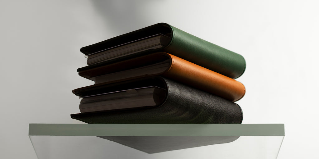 Conscious Vegan Leather Folios in colors Valley, Clay, and Mesa are stacked on a glass shelf. 