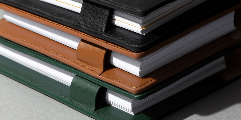 Conscious Vegan Leather Folios in colors Clay, Mesa, and Valley are stacked on a desk. 
