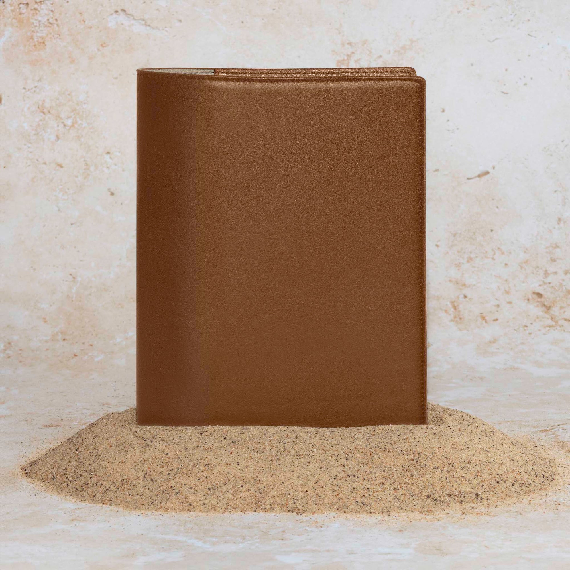 Folio displayed on a textured tan background, placed in a pile of sand. Color pictured is clay brown. 