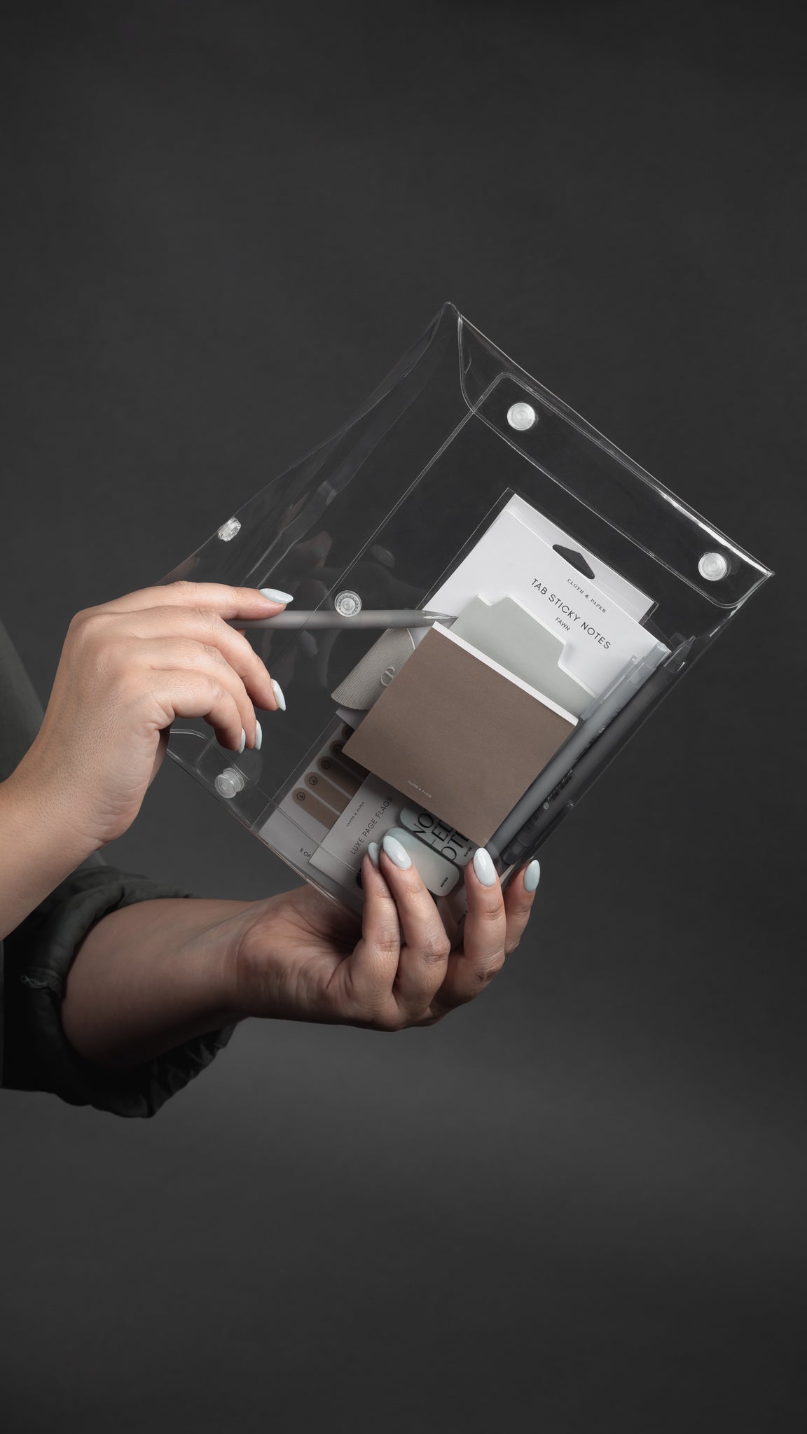 A hand holding a transparent pouch containing various stationery items, including sticky notes, pens, and a pencil.