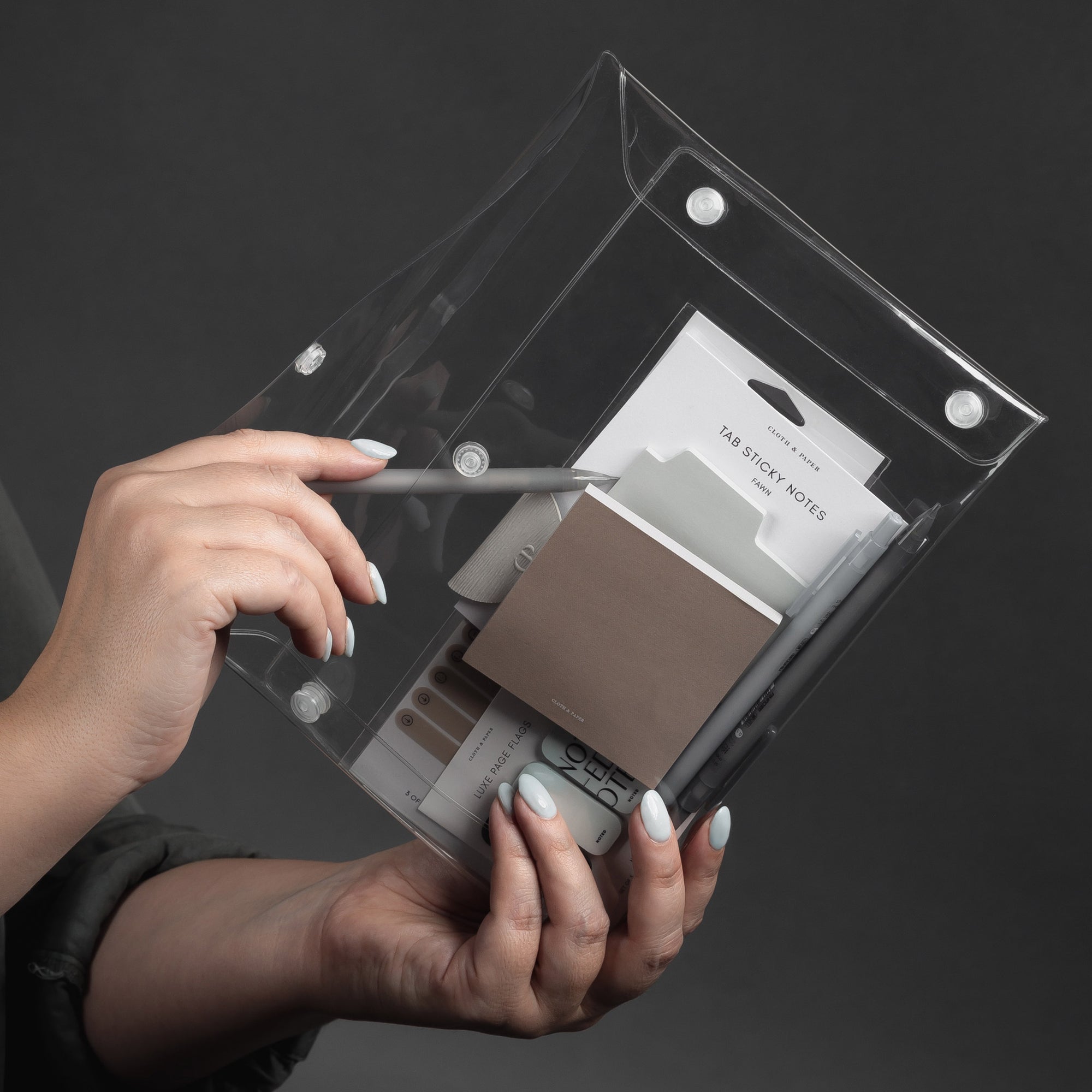 A hand holding a transparent pouch containing various stationery items, including sticky notes, pens, and a pencil.