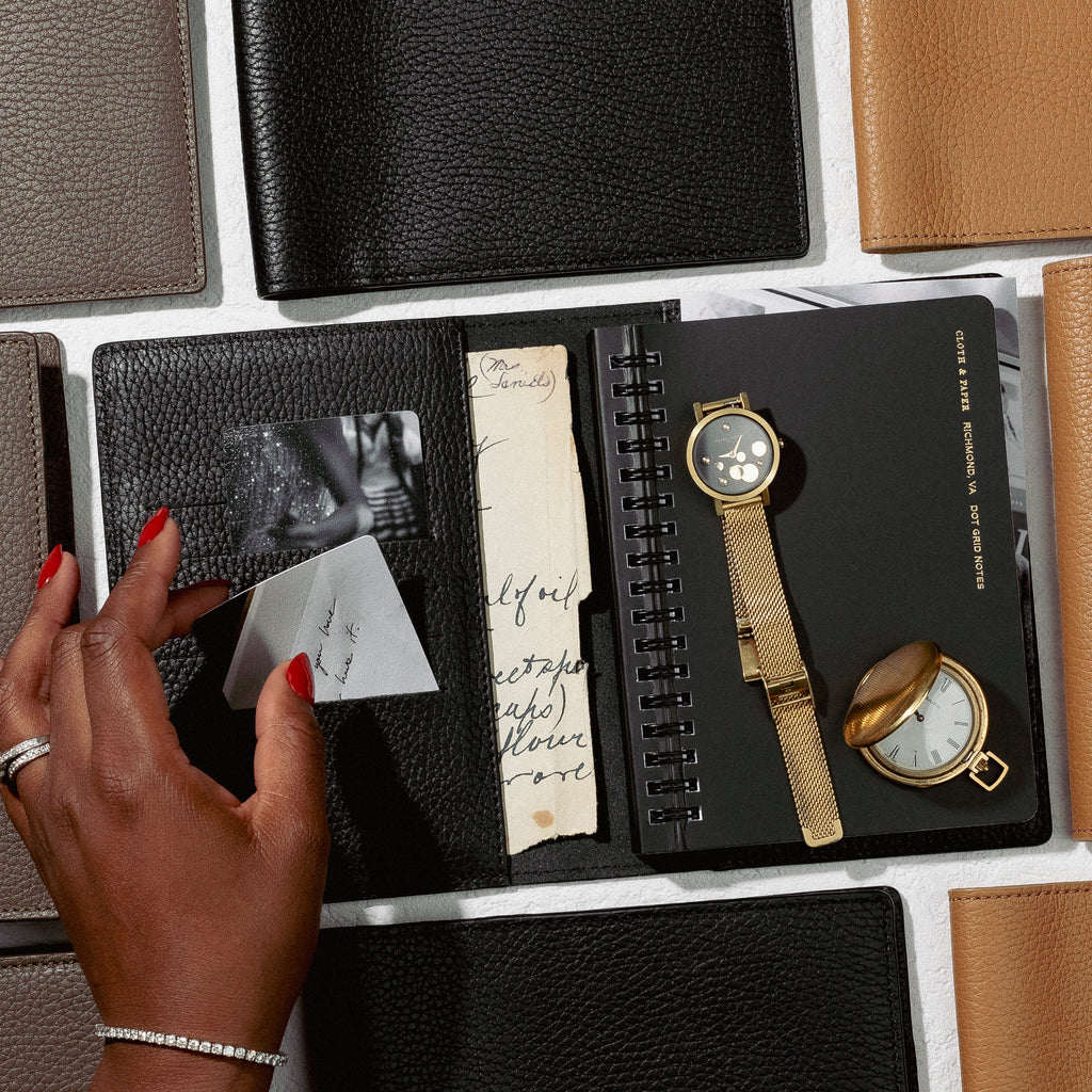 an image representing folios from our new Heirloom Leather Collection.  A hand is going through the contents of a planner with everyday accessories surrounding.