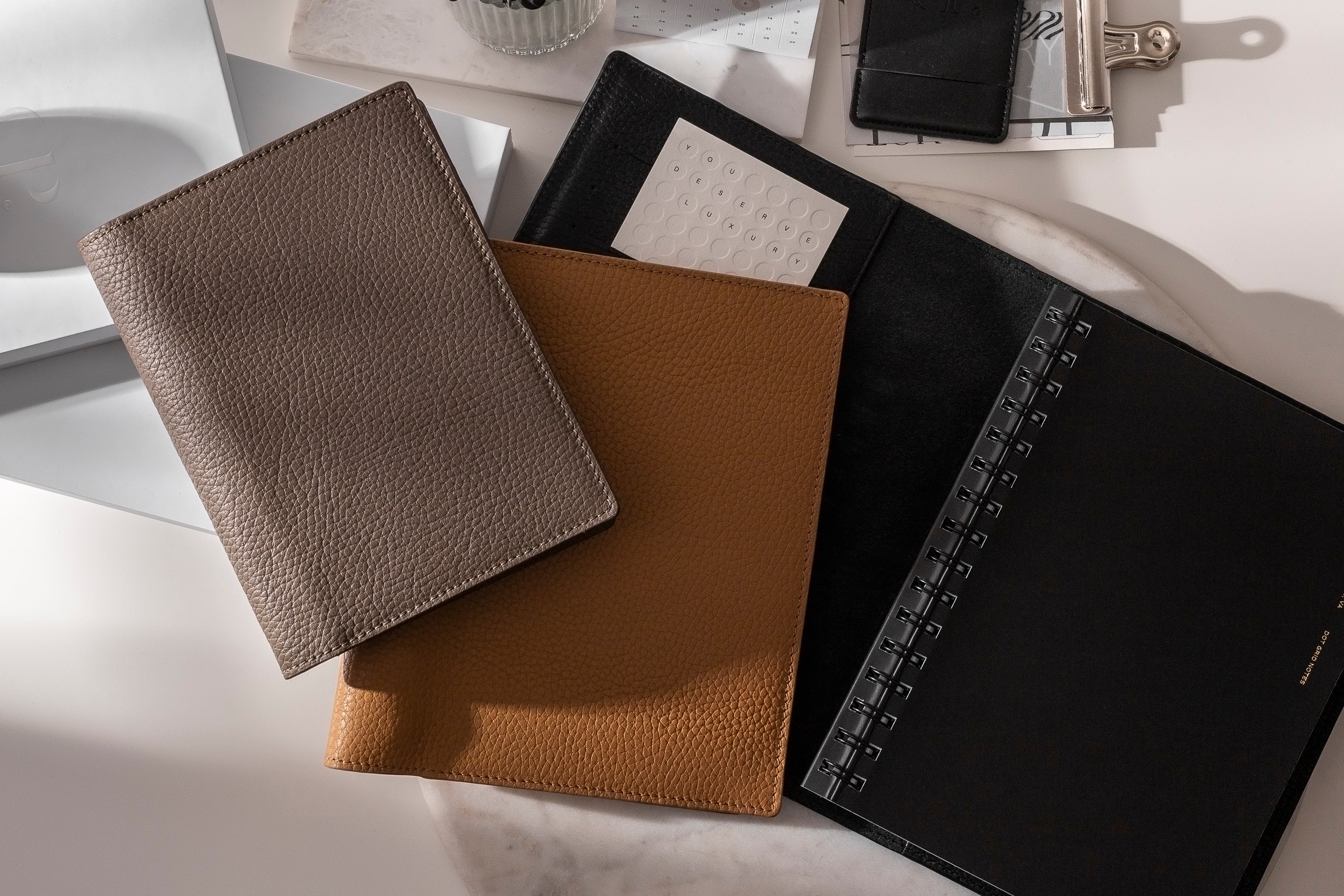 A few Heirloom Leather Folios are arranged on a white desk surrounded by other desk accessories.