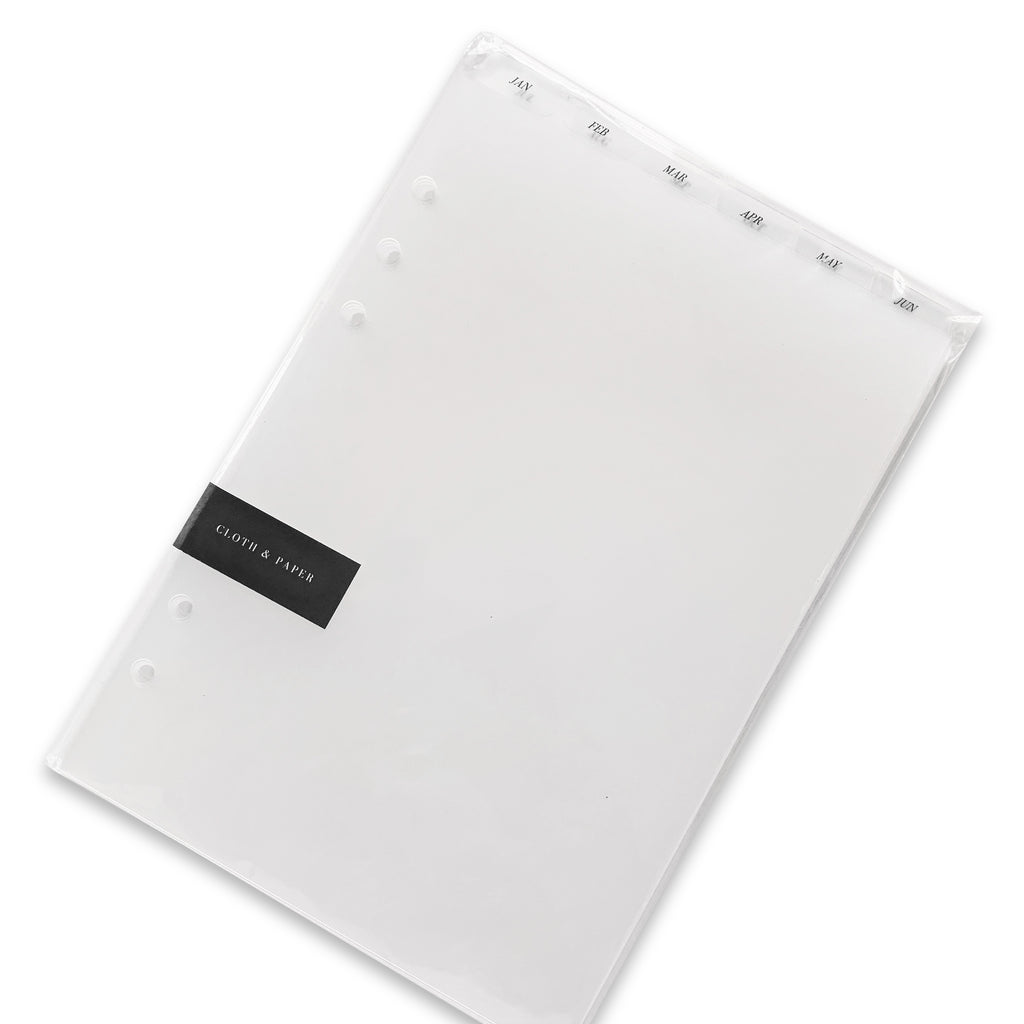 Monthly Top Tab Planner Dividers, Serif Style, Glass Plastic, Black Foil, Cloth and Paper. Dividers in their packaging tilted to the right on a white background.
