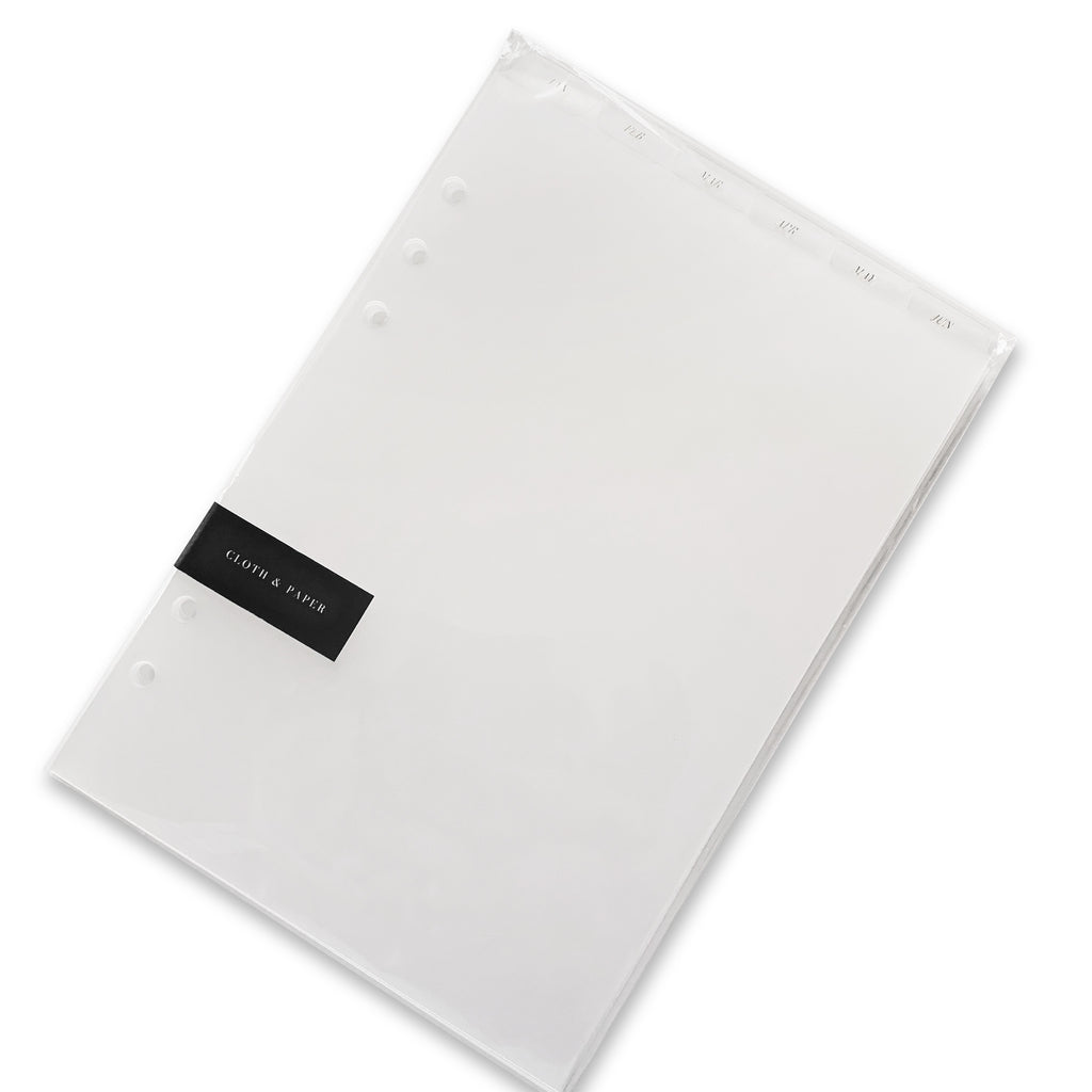 Monthly Top Tab Planner Dividers, Serif Style, Glass Plastic, Gold Foil, Cloth and Paper. Dividers in their packaging tilted to the right on a white background.