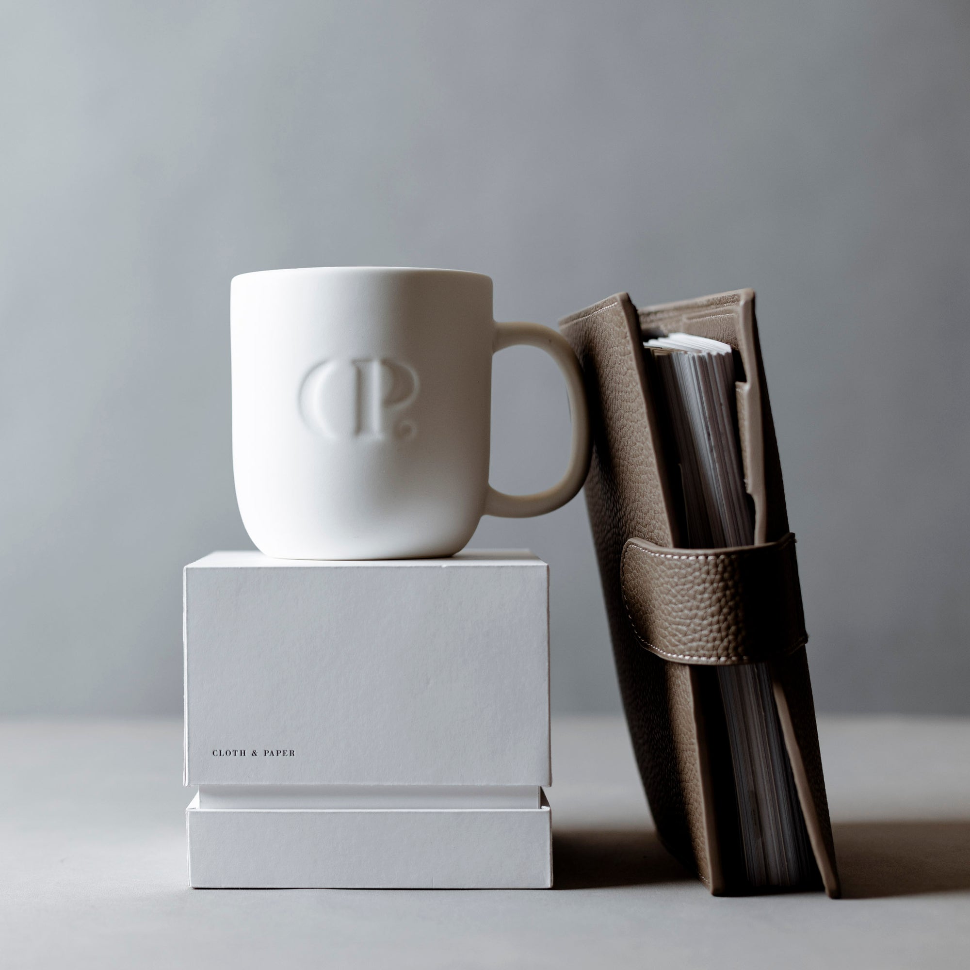 A Foundations Leather 6-Ring Agenda is leaning against a C&P Mug.  Both items are sitting on a white desk against a gray background.