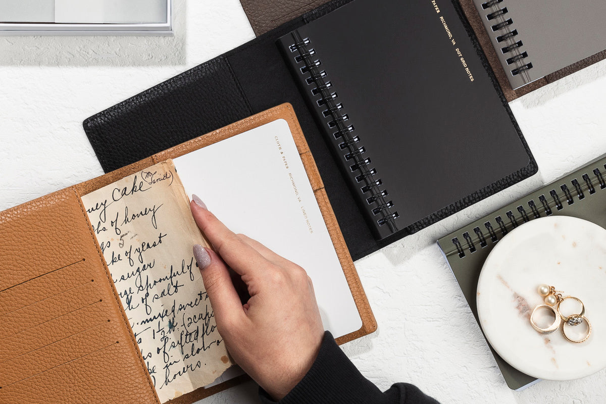 A few Heirloom Leather Folios are opened on a white desk.  A hand is adding some pages of notes to one of the inner pockets.