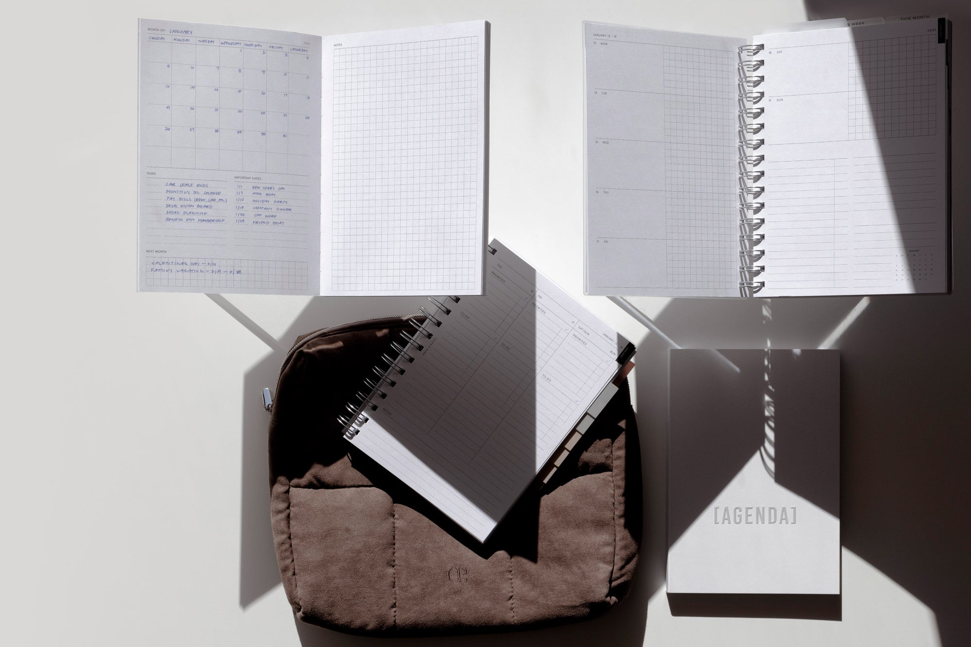 New Arrivals in Planners and Stationery are sitting on a desk with some shadows overhead.  These New Arrivals include the Undated Admin Perfect Bound Notebook, 2025 Spiral Bound Planners, and our 2025 Spiral Planner Single Purchase Subscription Box.