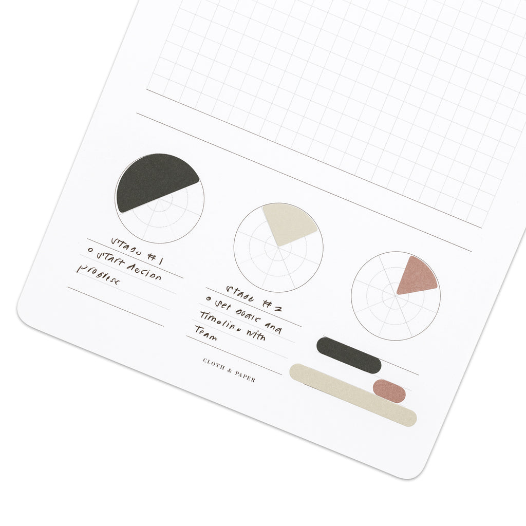 Stickers in use on a Progress Tracker A5 Notepad. One of each kind of sticker - pie chart and bar graph - are placed around the bottom of the notepad, and some example text is written underneath the leftmost and center printed pie charts.