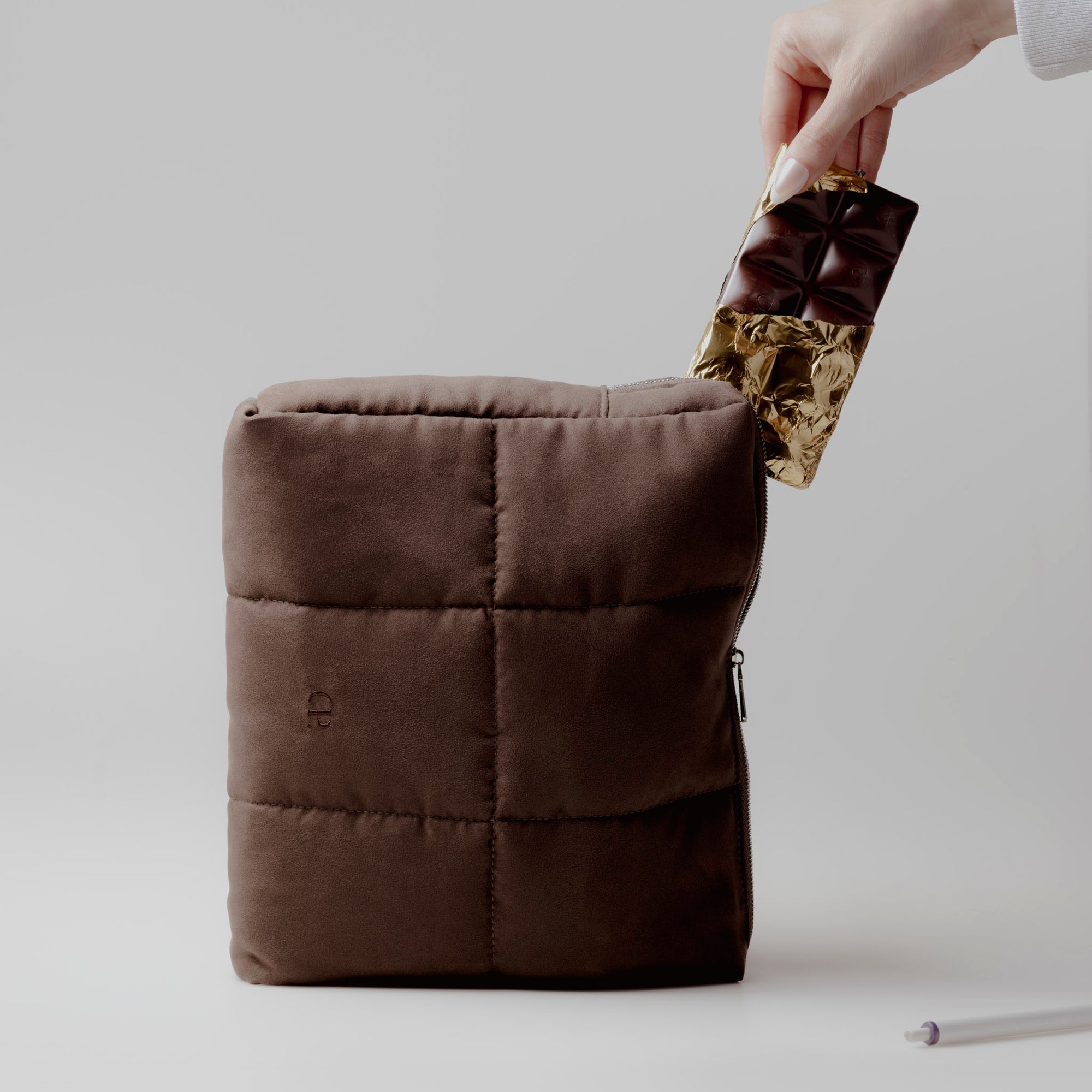 A hand is placing a chocolate bar inside our Puffer Pouch in color Café Noir.  The Puffer Pouch is standing up against a light gray background.