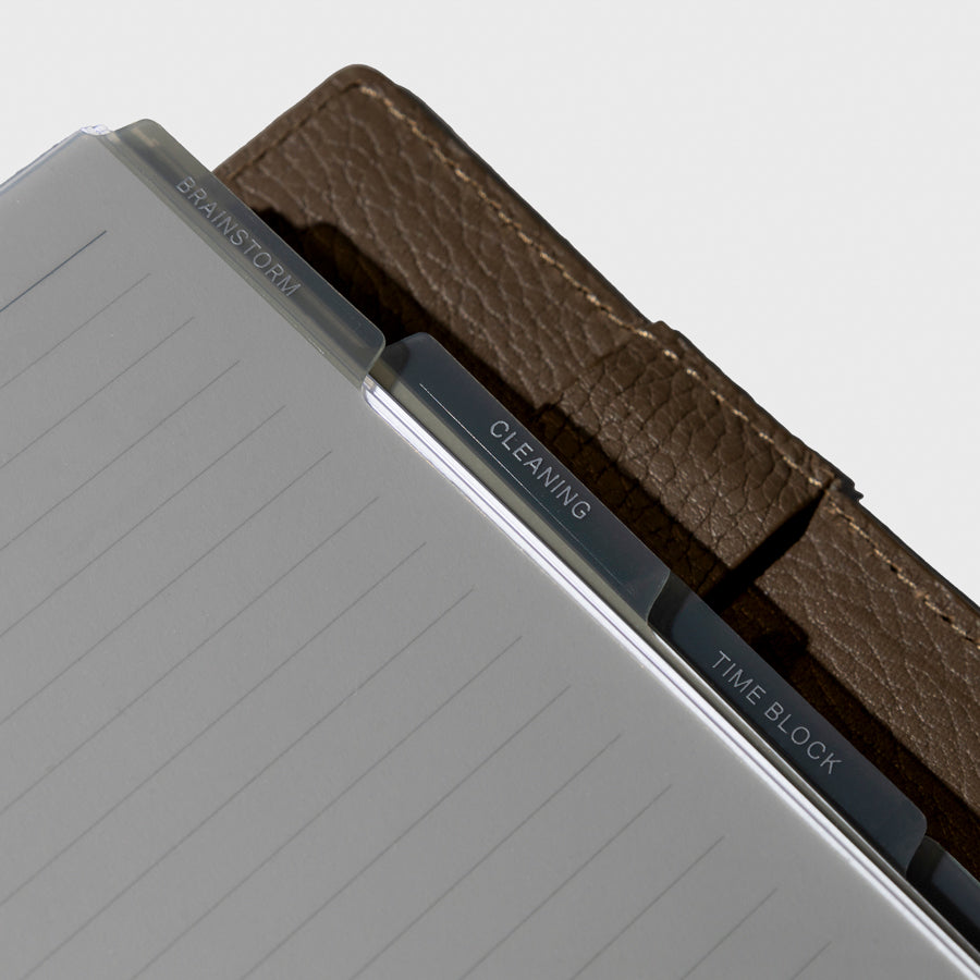 A close up image of the tinted gray dividers in use inside a brown leather planner. The divider tabs have labels on them that say brainstorm, cleaning  and time block