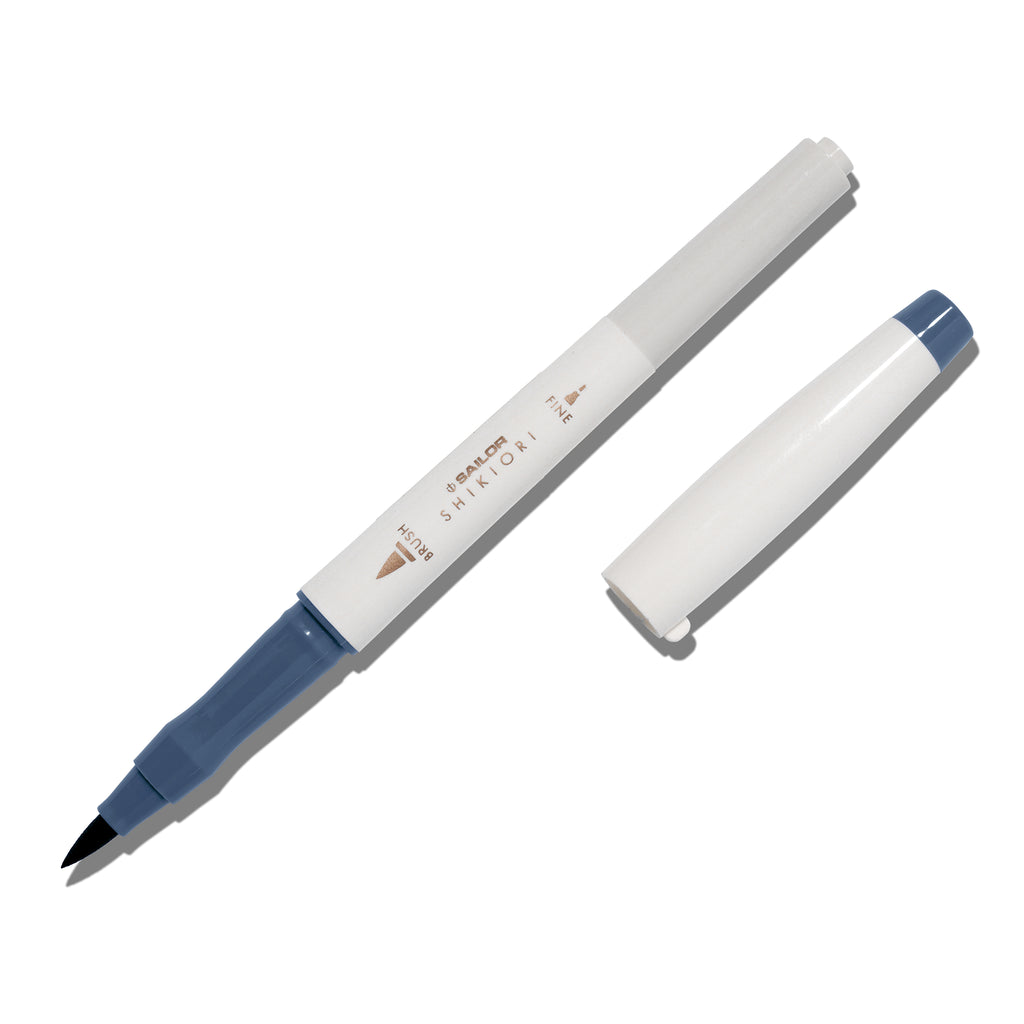 Brush pen displayed on a white background. It is uncapped with its brush nib exposed, and the cap lying parallel to the marker. Color pictured is Shimoyo. 