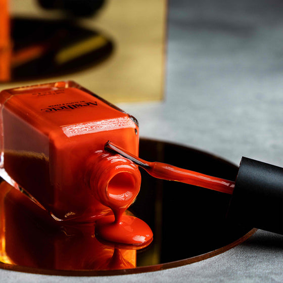 Aesthete Nail Polish, Cloth and Paper. An opened bottle of the nail polish, which is bright red, rests on a mirrored display. The applicator brush rests on the neck of the nail polish bottle.