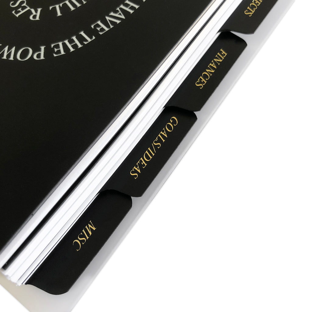CEO Side Tab Planner Dividers, Black Plastic, Gold Foil, Cloth and Paper. Close up of dividers in use in a planner tilted to the right on a white background.