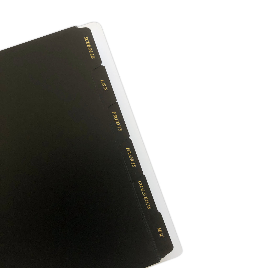 CEO Side Tab Planner Dividers, Black Plastic, Gold Foil, Cloth and Paper. Close up of dividers in use in a planner tilted to the left on a white background.