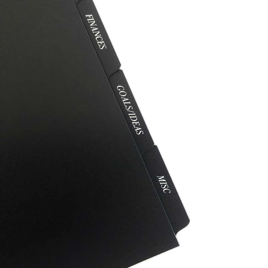 CEO Side Tab Planner Dividers, Black Plastic, White Foil, Cloth and Paper. Close up of dividers in use in a planner tilted to the left on a white background.
