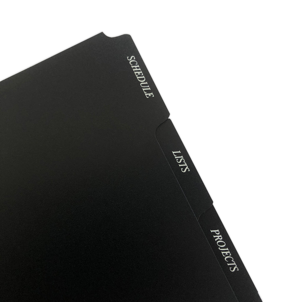 CEO Side Tab Planner Dividers, Black Plastic, White Foil, Cloth and Paper. Close up of dividers in use in a planner tilted to the left on a white background.