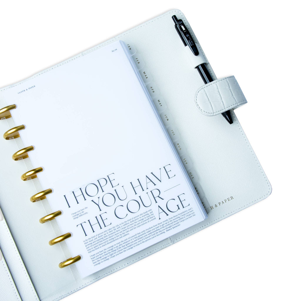 Courage Planner Dashboard styled inside a discbound planner in a white leather agenda cover. The discbound planner has gold rings, and there is black pen tucked into the agenda cover's pen loop.