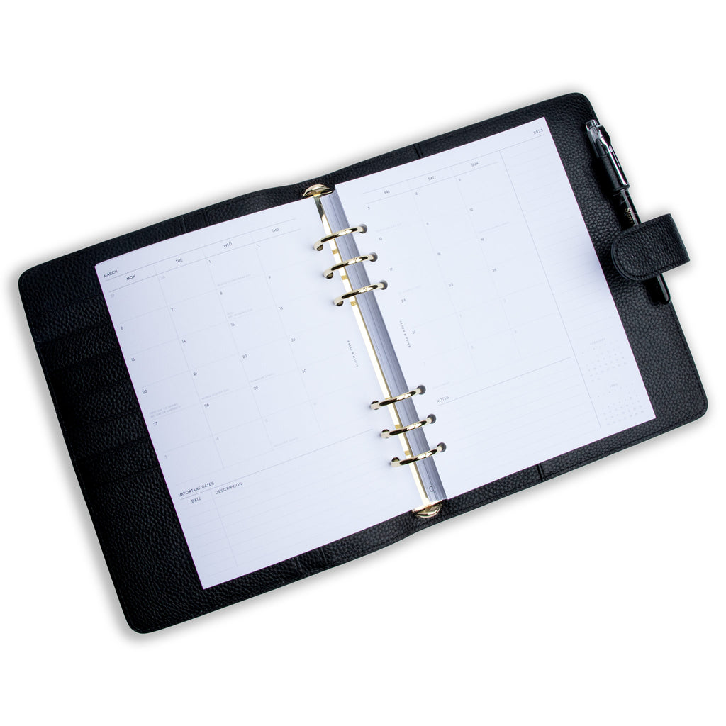 2023 Dated Planner Inserts, Monthly, Monday Start, Cloth and Paper. Inserts in use inside a black leather agenda with silver rings on a white background. A pen rests in the agenda's pen loop.