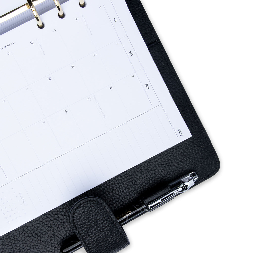 2023 Dated Planner Inserts, Monthly, Monday Start, Cloth and Paper. Close up of inserts in use inside a black leather agenda with silver rings. A black pen is inside the agenda's pen loop.
