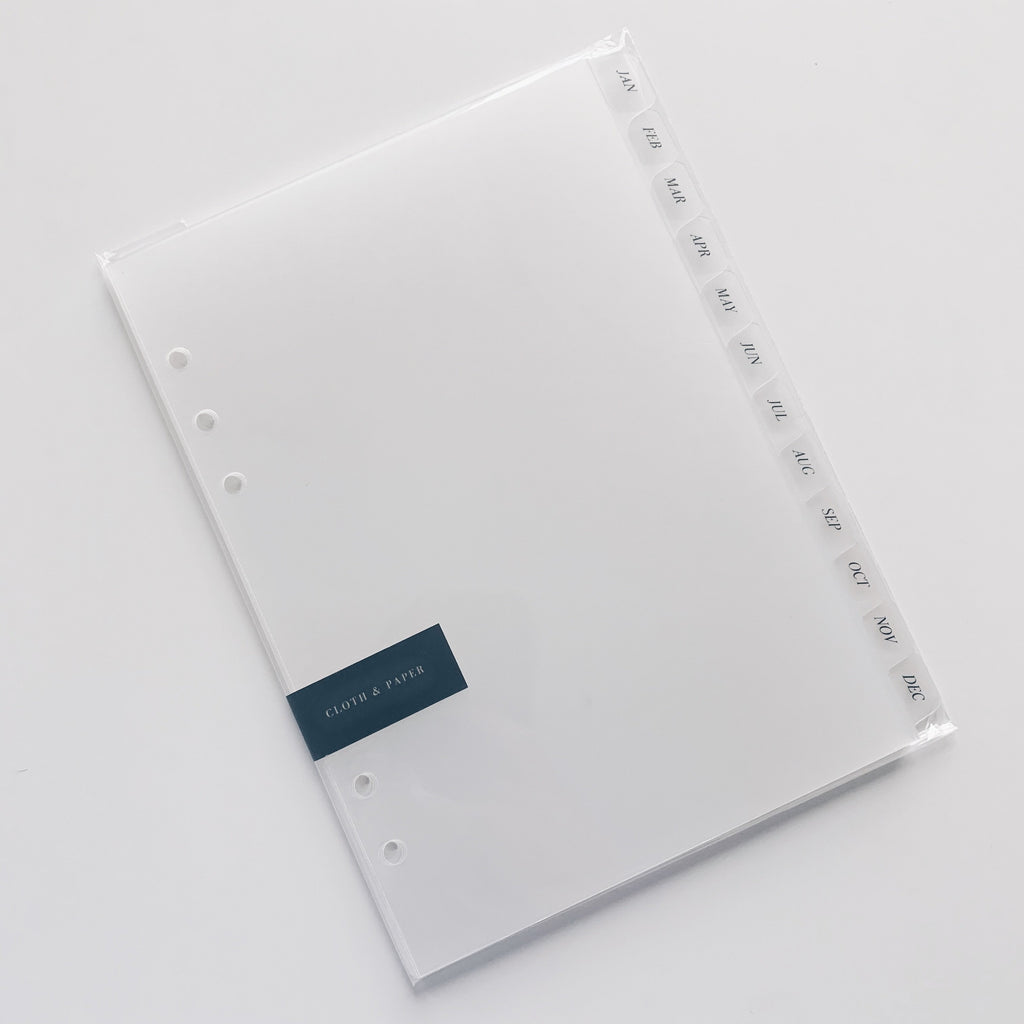 Monthly Side Tab Planner Dividers, Glass Plastic, Black Foil, Cloth and Paper. Dividers in their packaging on a white background tilted to the left.