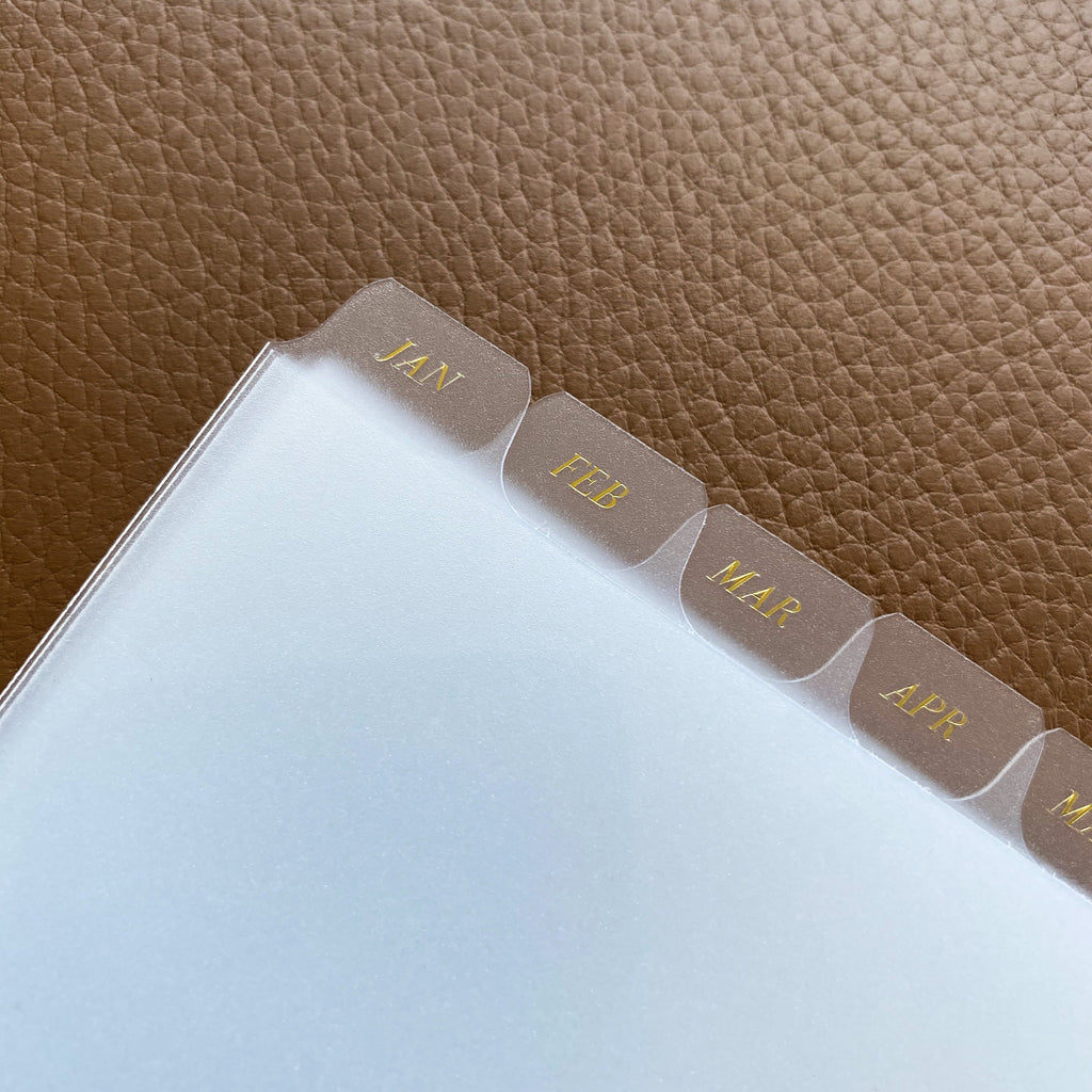 Monthly Side Tab Planner Dividers, Glass Plastic, Gold Foil, Cloth and Paper. Close up image of dividers on a brown leather background.