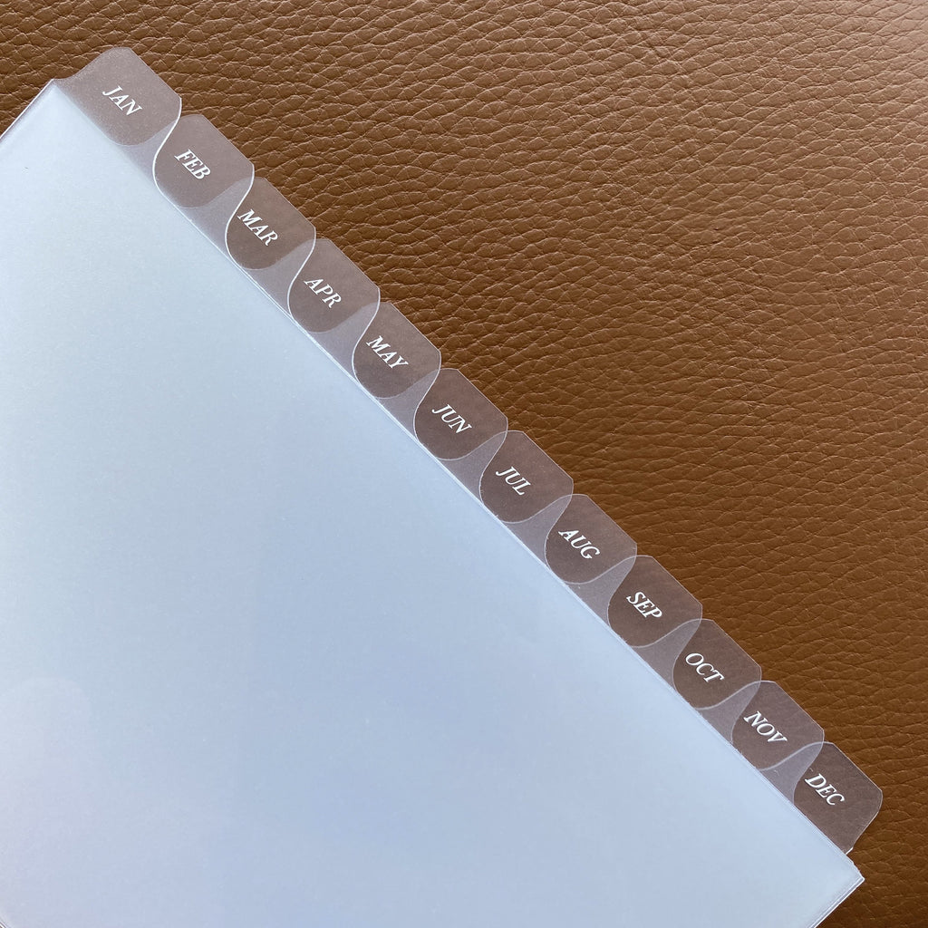 Monthly Side Tab Planner Dividers, Glass Plastic, White Foil, Cloth and Paper. Close up image of dividers on a brown leather background.