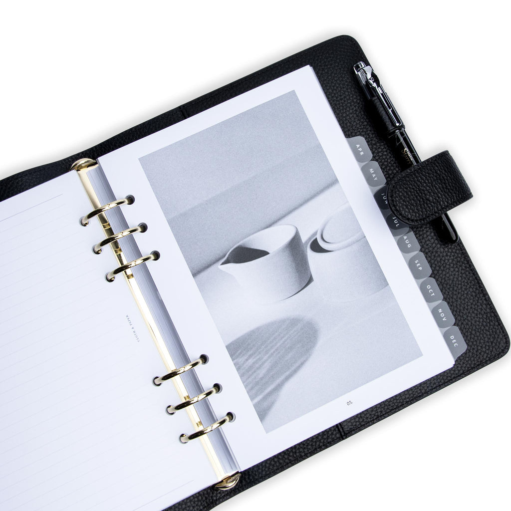 2023 Dated Planner Inserts, Vertical Weekly Lined, Monday Start, Cloth and Paper. Insert in use inside a black leather agenda with silver rings and clear monthly divider tabs on a white background. A black pen rests inside the agenda's pen loop.