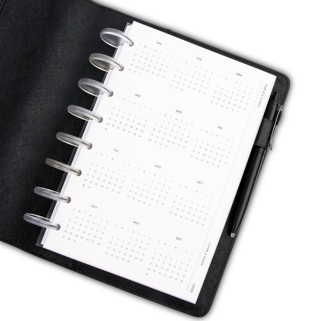 2023 Year at a Glance Dashboard, Cloth and Paper. Dashboard in use inside of a black leather agenda cover with clear discs. A pen rests in the agenda's pen loop.