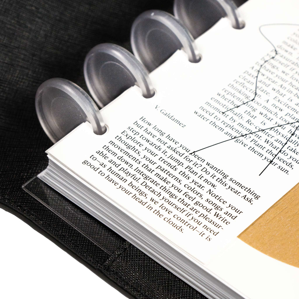 Close up on bottom left corner of dashboard styled inside a discbound planner and black leather planner cover. Text from prose written by V. Galdamez is in focus.